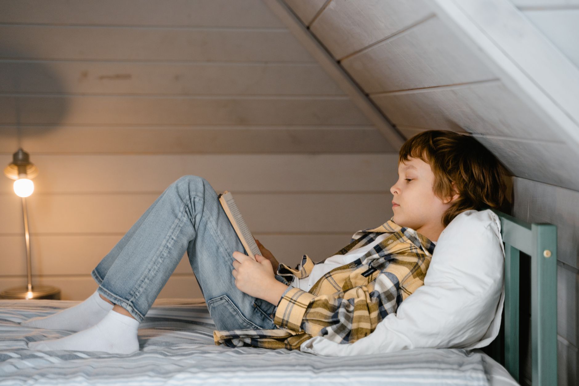 A young boy in plaid relaxes with a book in a cozy attic bedroom setting.