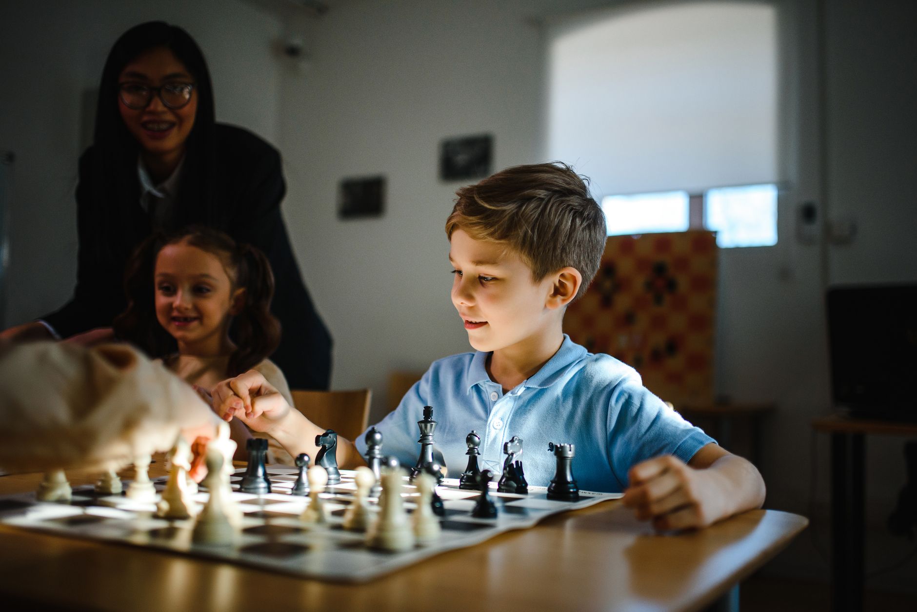 Young boy and girl playing chess in a classroom under teacher's guidance.