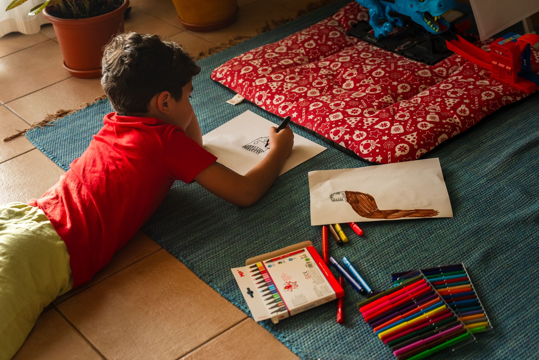 A young boy creatively drawing pictures while lying on the floor with colorful markers.