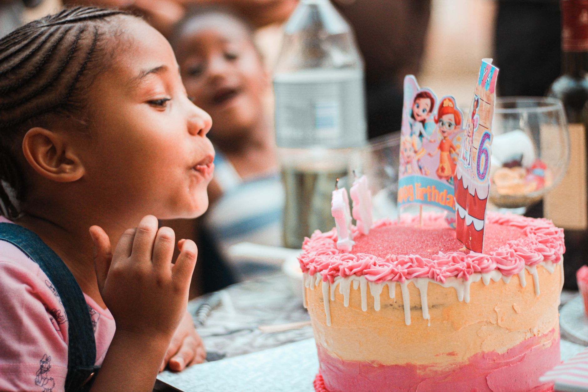 Side view of crop adorable little black girl with Afro braids blowing out candles on birthday cake during party