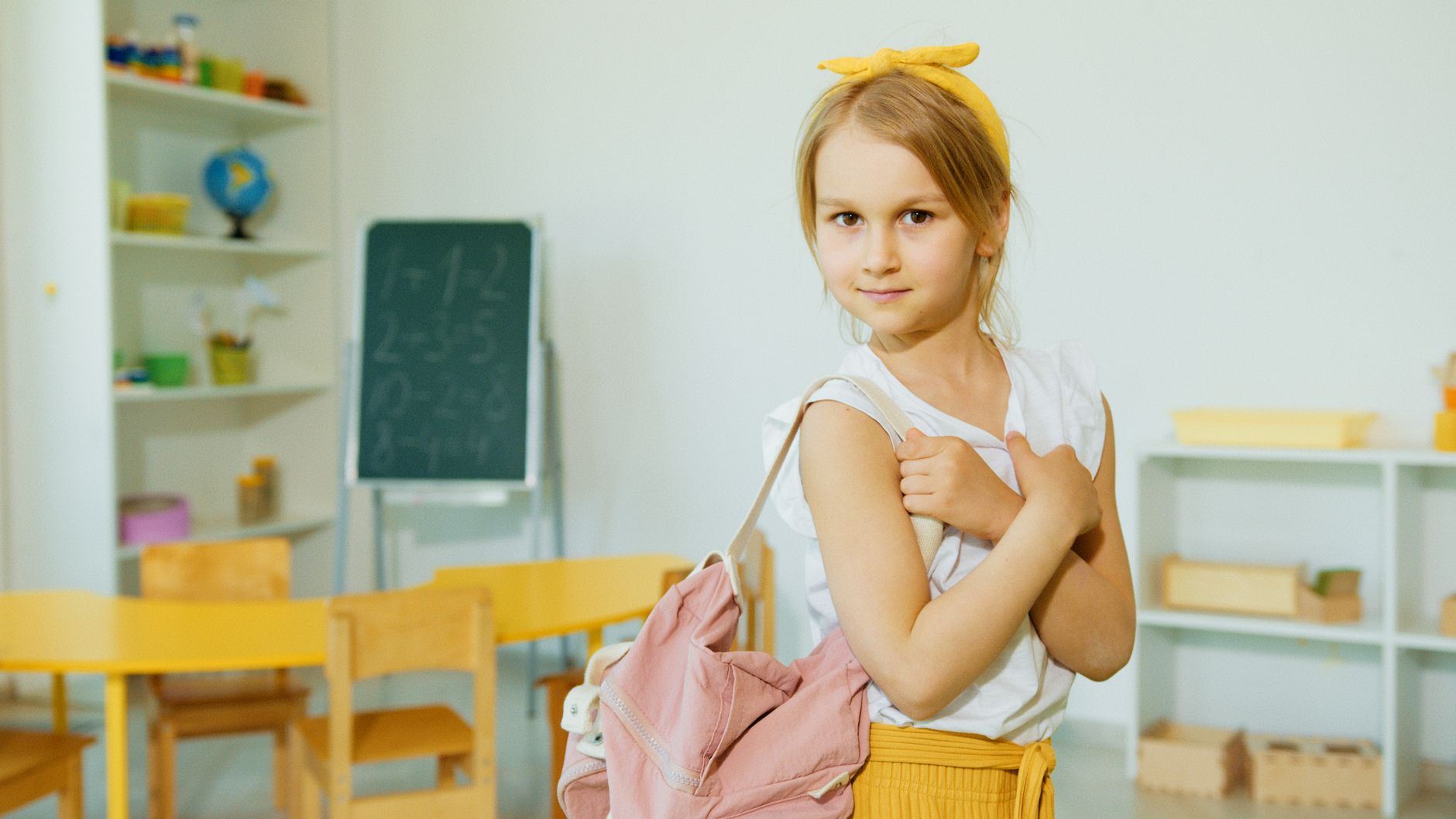 A young girl in a classroom holding a backpack, surrounded by educational materials.