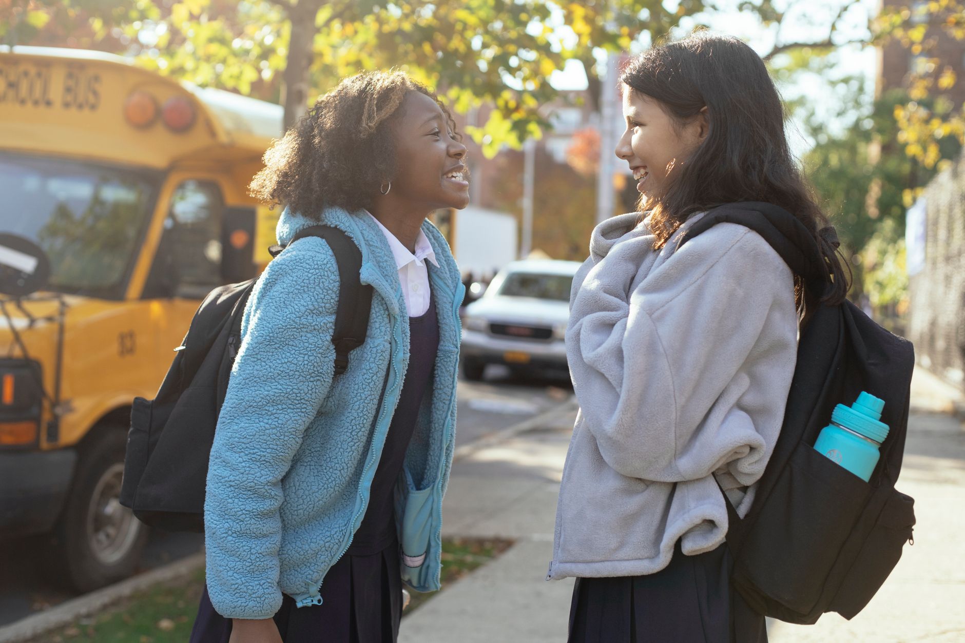 Side view happy multiracial schoolgirls in warm jackets with backpacks standing on street near school bus and talking while looking at each other with smile
