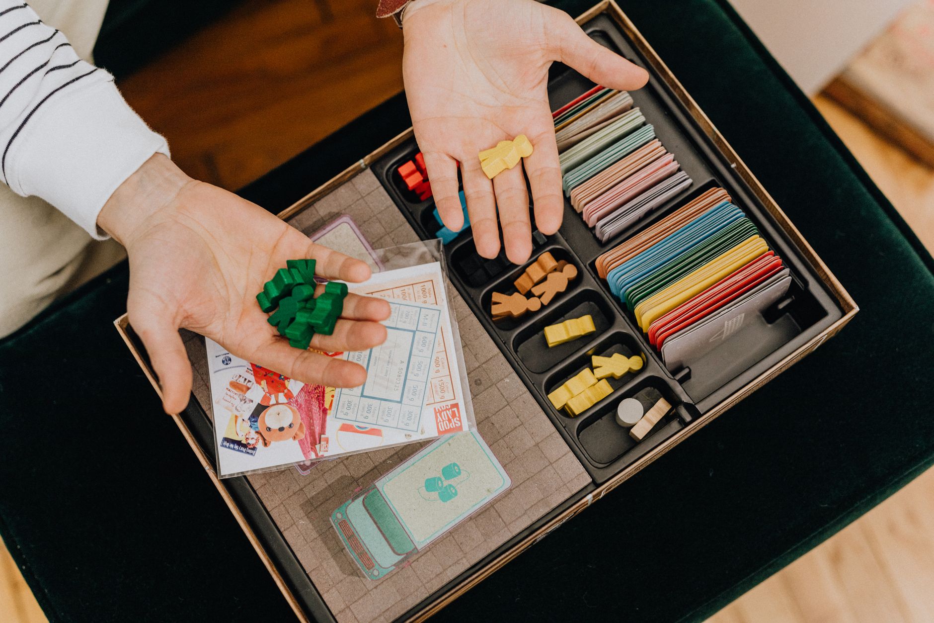 Colorful board game components being held in hands above a game box, top view.