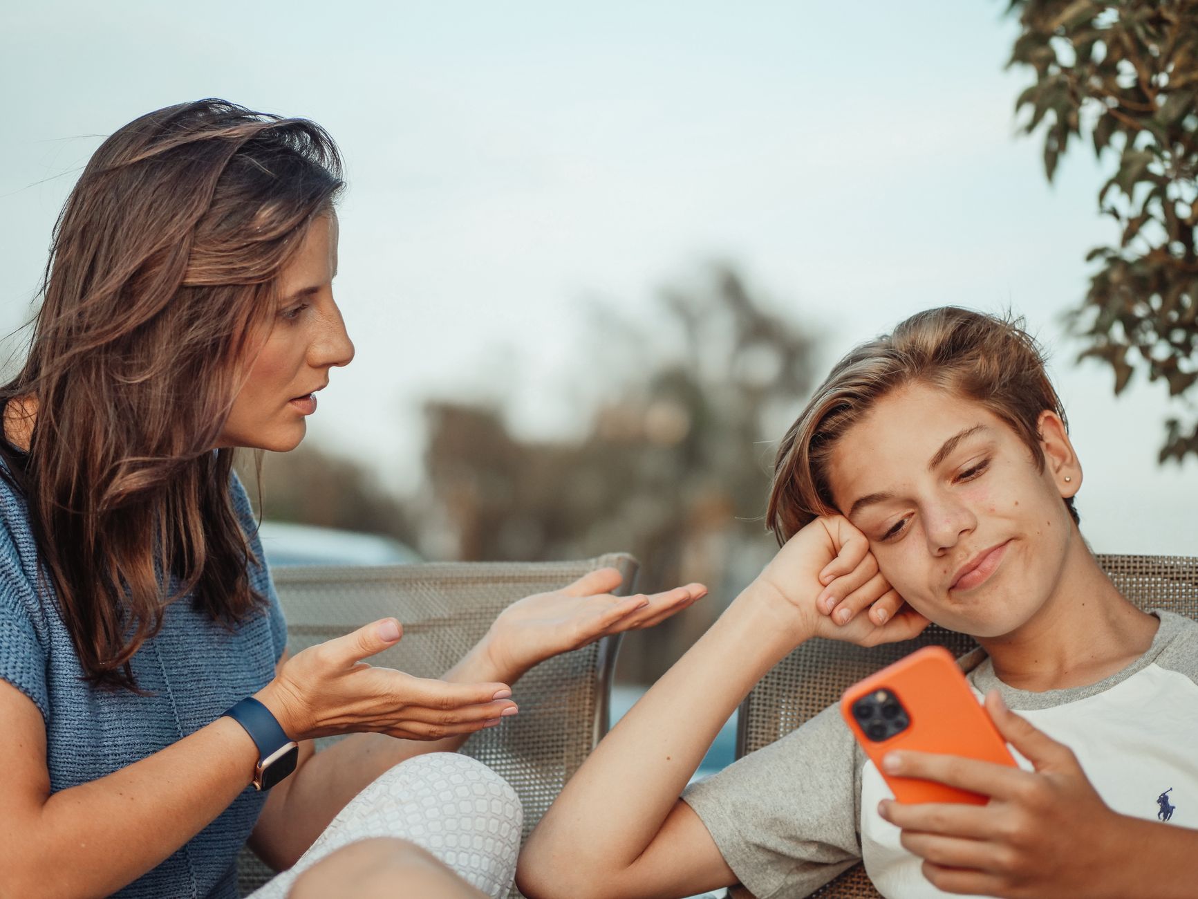 A mother engaging with her teenage son holding a smartphone outdoors.