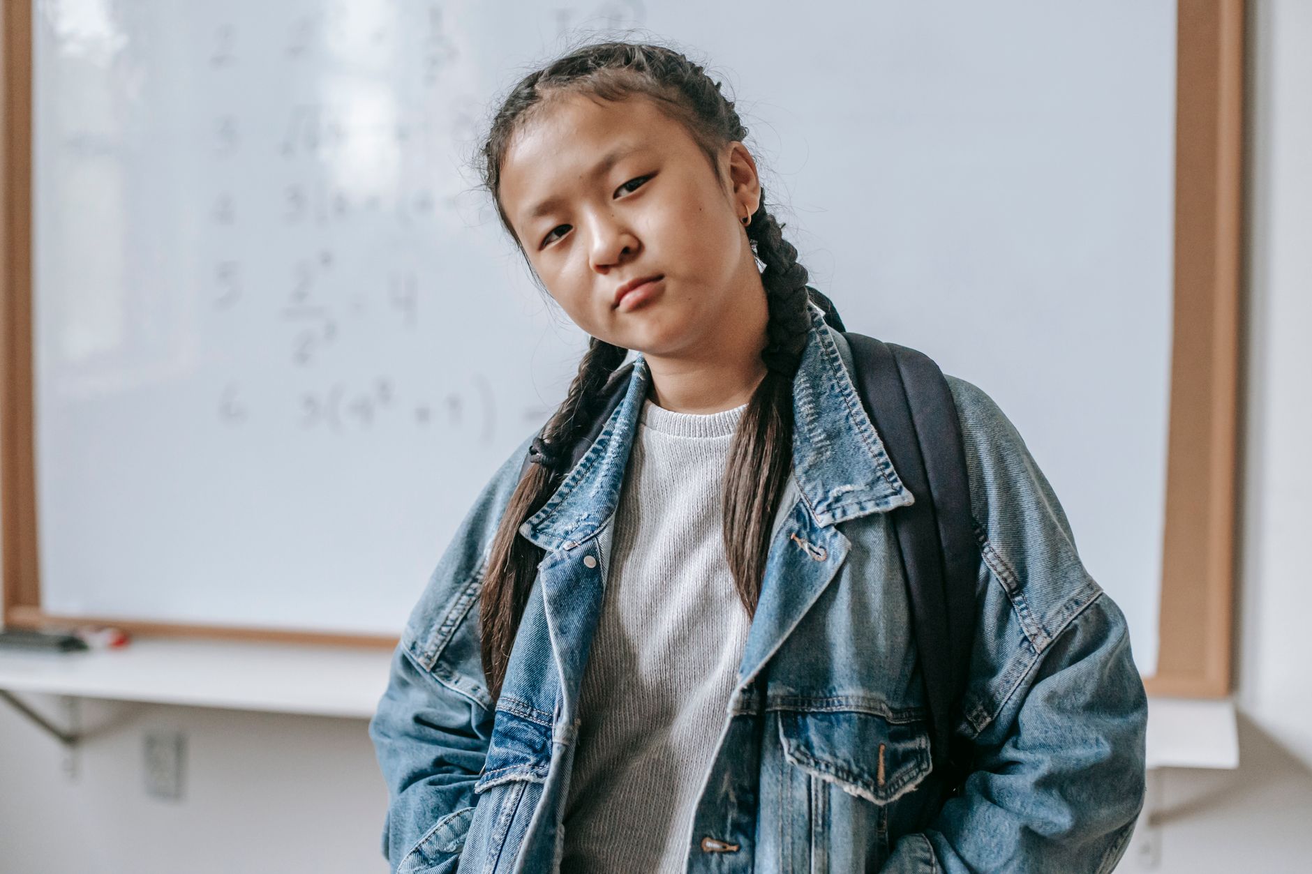 Concentrated Asian girl in denim jacket with backpack and hands in pockets standing in classroom against whiteboard and looking at camera