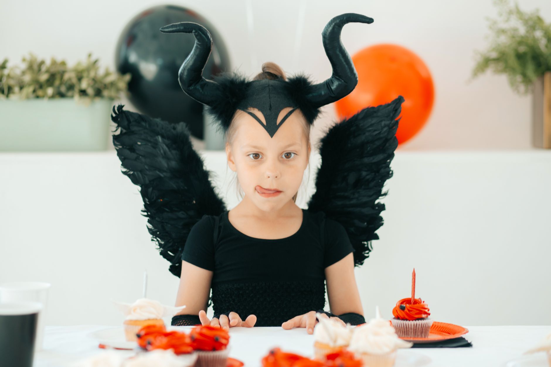 A girl in a devil costume enjoying cupcakes at a Halloween-themed celebration indoors.