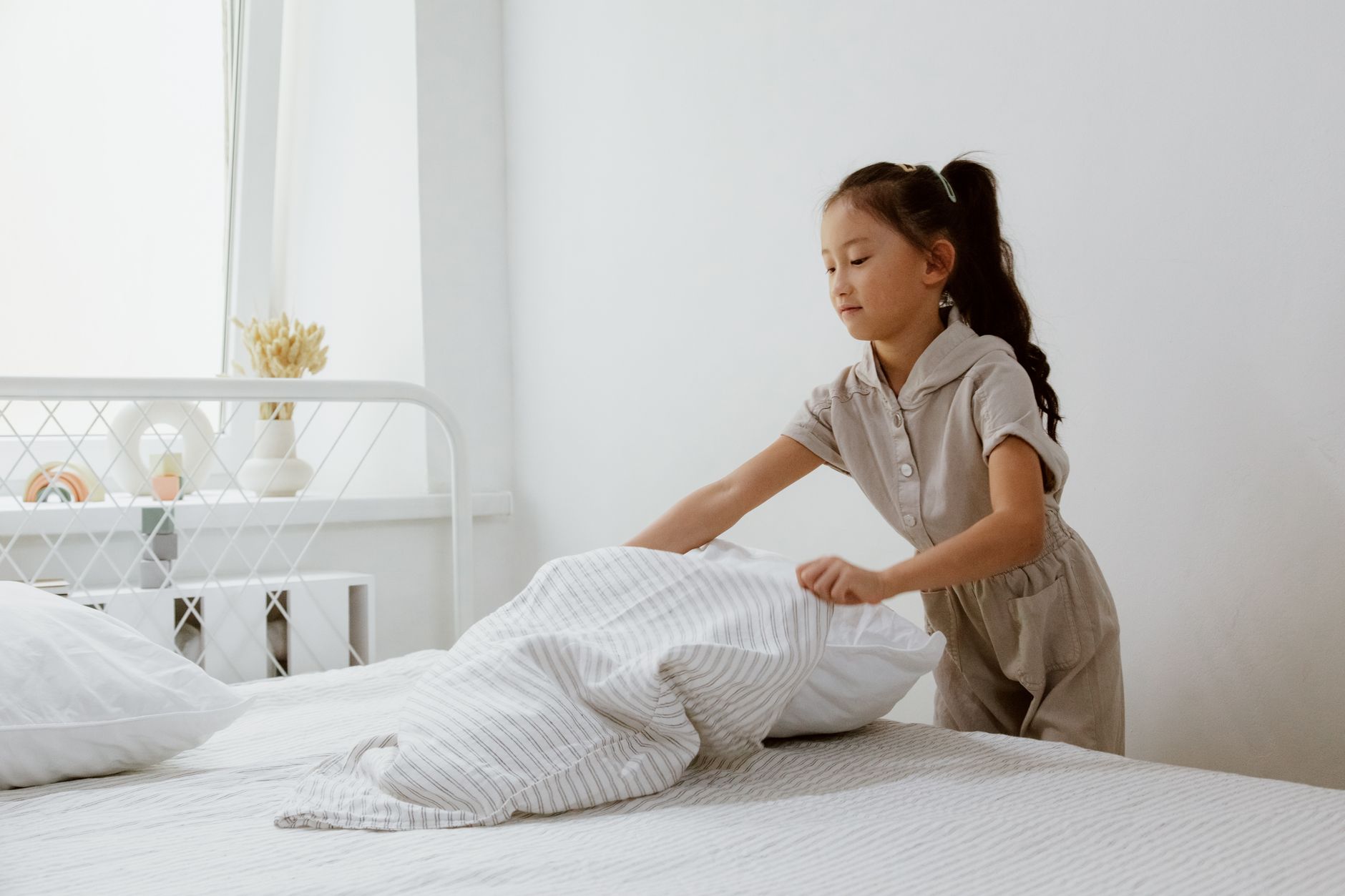 A girl making her bed in a bright, cozy bedroom with a window view.