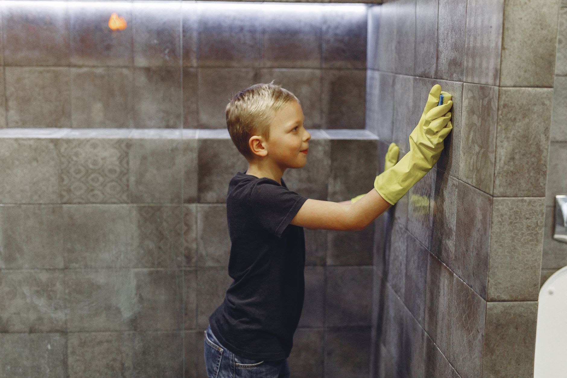 Side view of kid with blond hair in yellow rubber gloves cleaning tile wall in bathroom