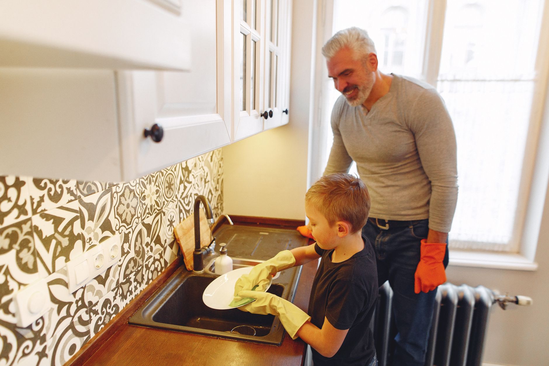 From above of grey haired father with beard teaching son dishwashing at kitchen