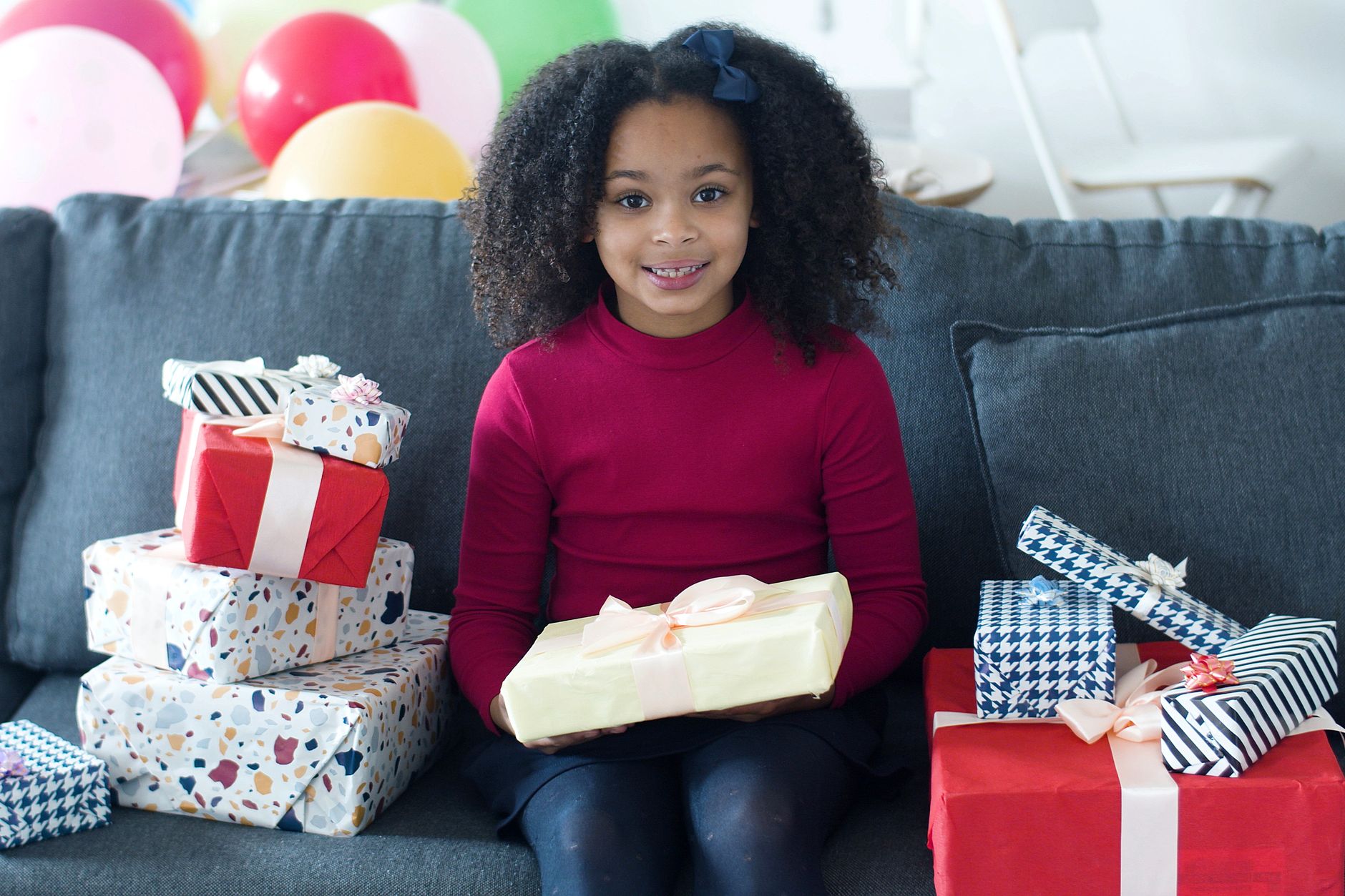 Young girl sitting on a sofa surrounded by beautifully wrapped presents, smiling joyfully for her birthday celebration.
