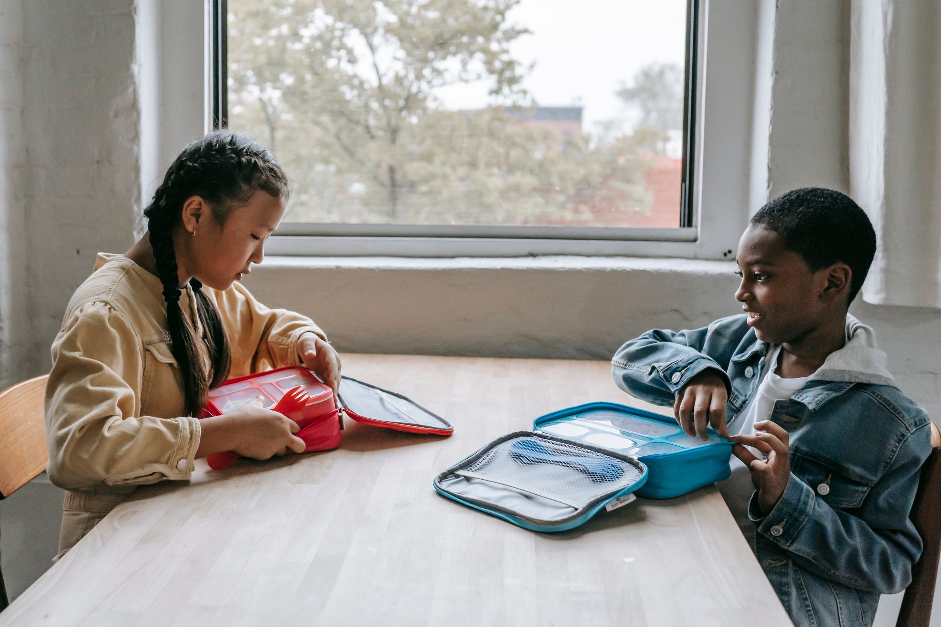 Side view of diverse children in casual clothes sitting at table with lunch boxes during break in school
