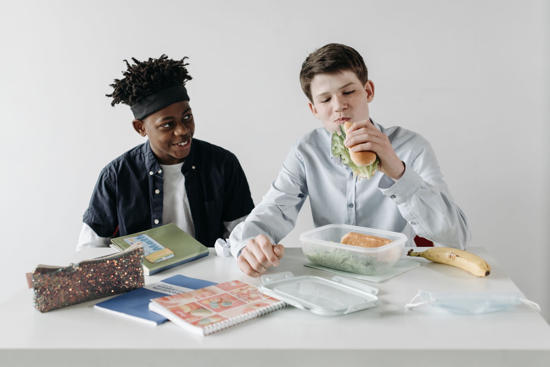 Two schoolboys enjoy lunch together at a white desk, featuring sandwiches and healthy snacks.