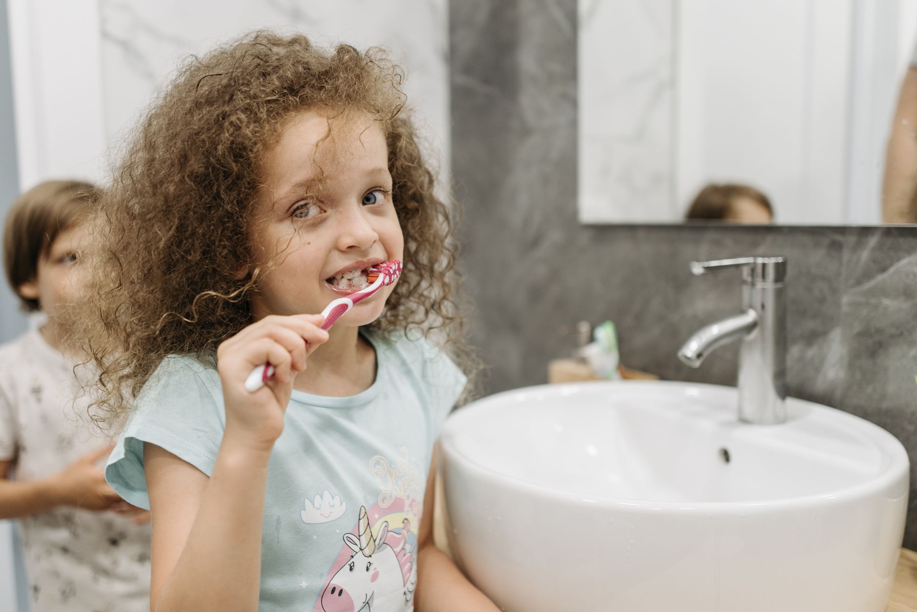 A young girl with curly hair brushes her teeth at a sink, promoting dental hygiene.