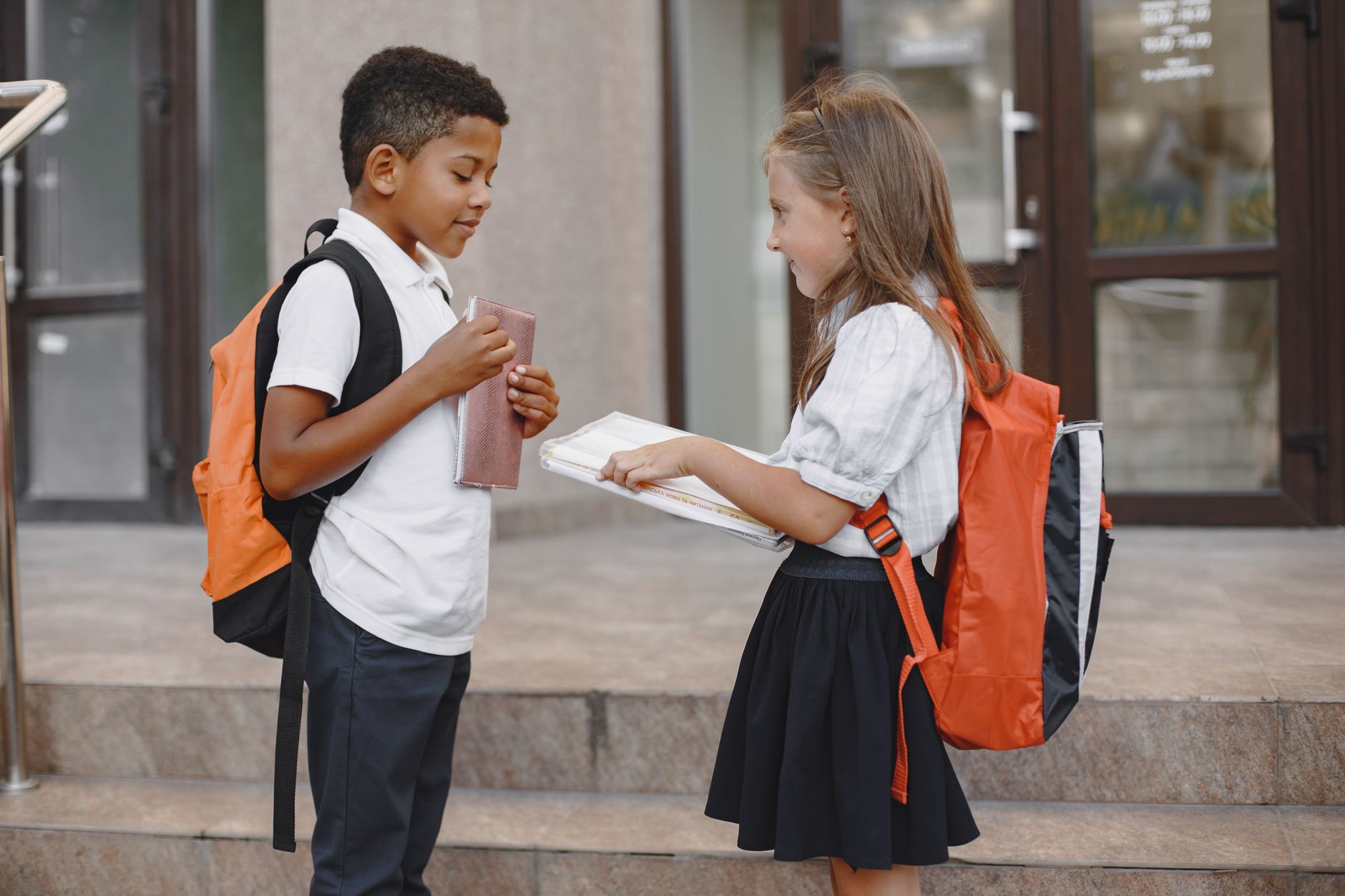 Two children with backpacks exchanging books outside a school building.