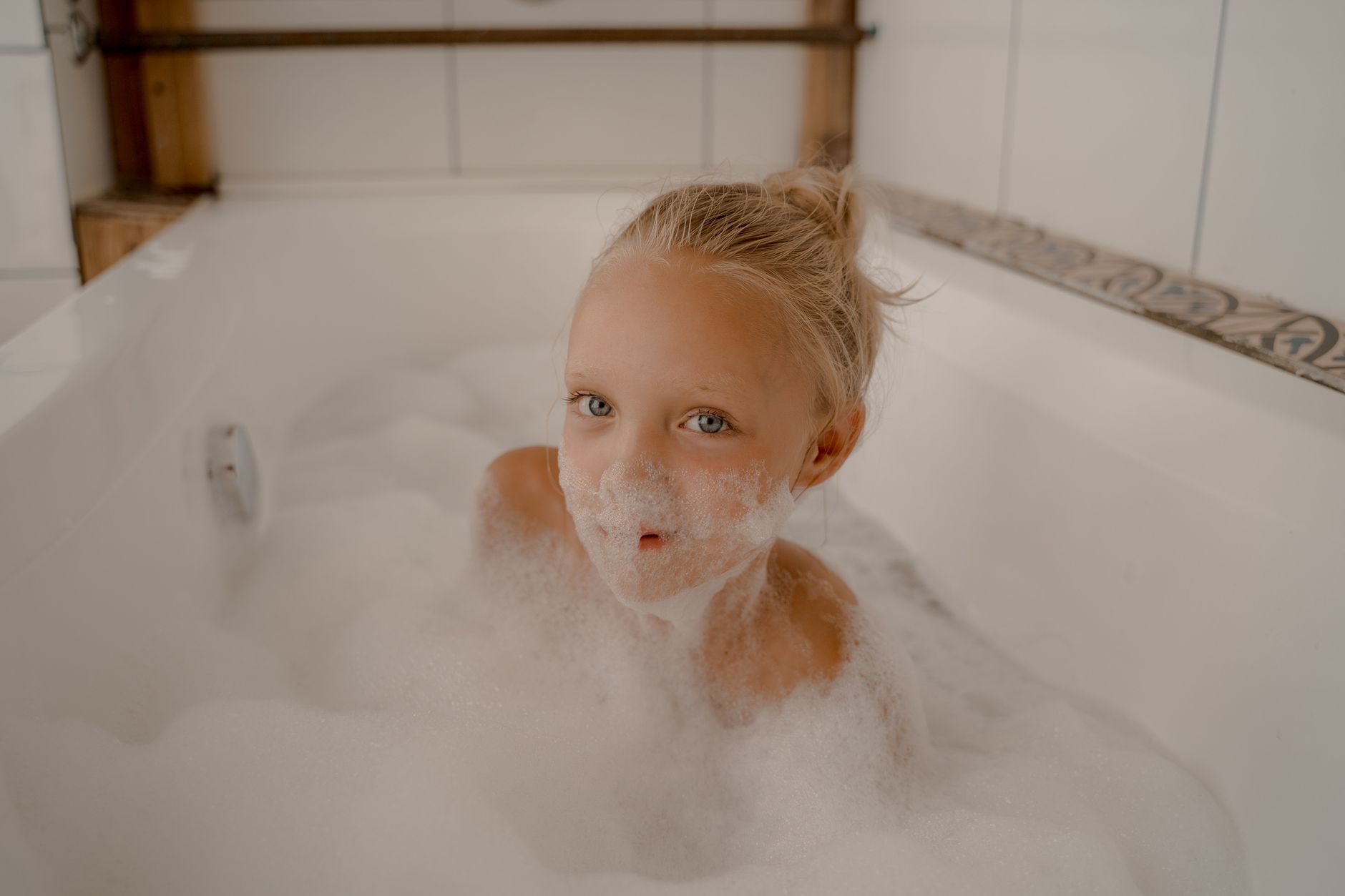 A young girl with blond hair enjoys a playful bubble bath in a white bathtub indoors.